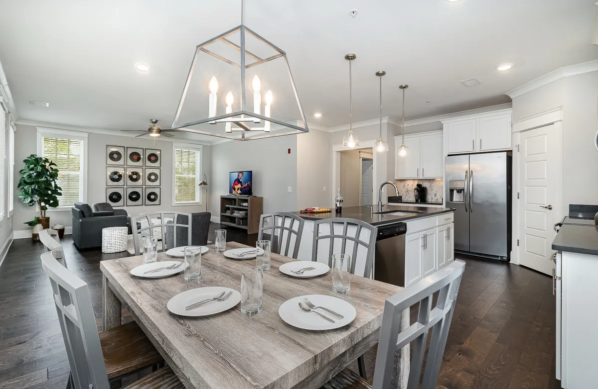This image presents an open-plan space that seamlessly blends a dining table set for a meal, a kitchen island, and a comfortable living area with a couch and TV. Decorative wall art and tasteful lighting contribute to a warm, welcoming atmosphere. The area is filled with natural light, highlighting the inviting communal spaces perfect for gathering, relaxing, and entertaining.