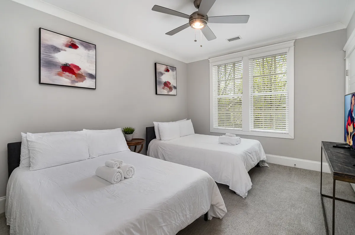 A calm and tranquil bedroom with two double beds dressed in crisp white linens, decorative wall art, and a window letting in abundant natural light. The setting is ideal for families or groups seeking comfort and privacy.