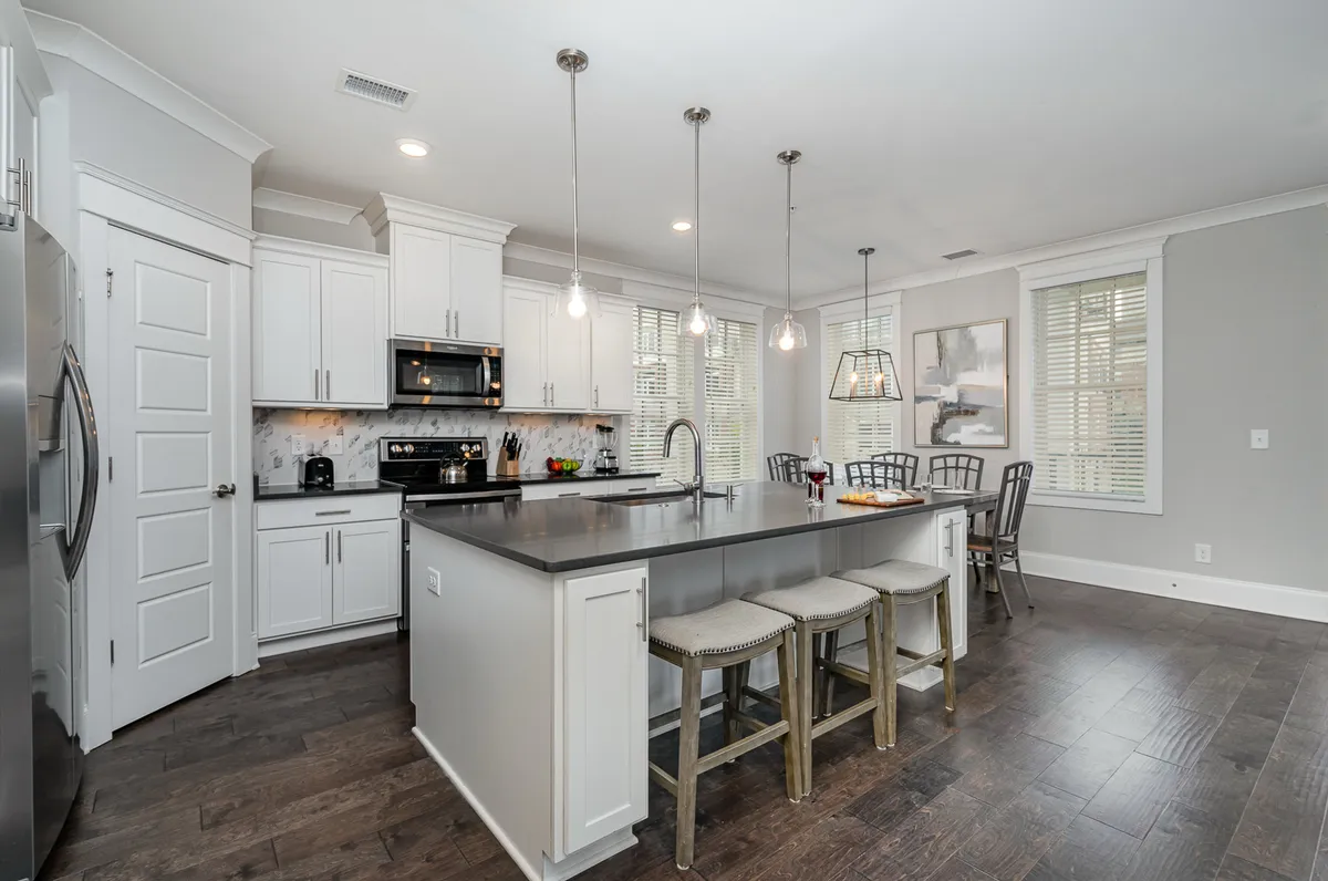 A vibrant, sunlit kitchen with a large island countertop, pendant lighting, modern appliances, and a spacious adjacent dining area. The living space is visible in the background, further reinforcing the open-concept design. The inviting setup is ready for shared meals, cooking together, or socializing over coffee.