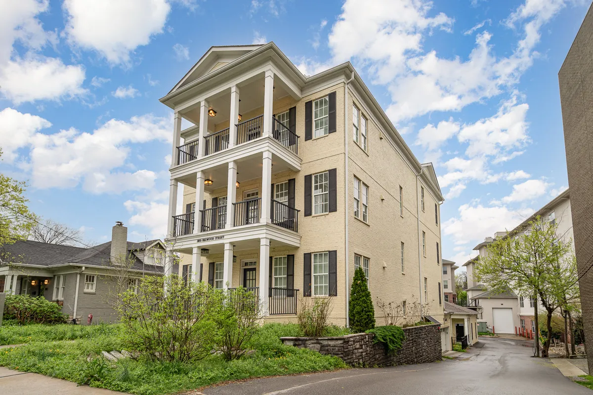 The image features the three-story exterior of the Willow property, showcasing balconies with sleek black railings and lush greenery in the foreground. The detailed brickwork and inviting facade evoke a sense of comfort and welcome.