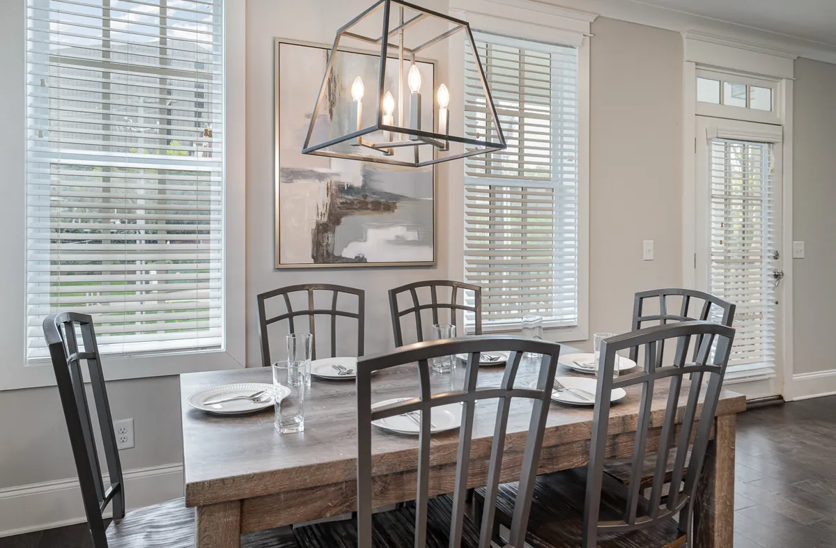 The bright and inviting dining space features a large table, elegant chandelier, and artwork by sunlit windows, creating a sense of comfort and warmth immediately upon entering the home.