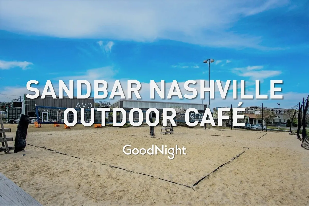 An animated exterior scene featuring an outdoor café with prominent signage, a sand volleyball court, and background buildings. The space buzzes with energy, combining socializing, dining, and recreation, all in a laid-back, pet-accessible atmosphere.